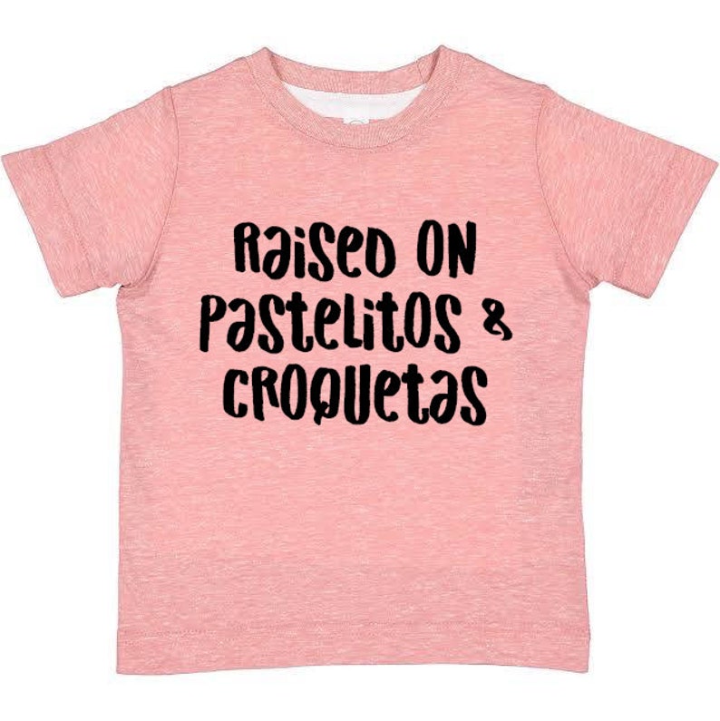 Op de afbeelding: Roze T-shirt met korte mouwen met de tekst "Raised on Pastelitos & Croquetas" in zwart.