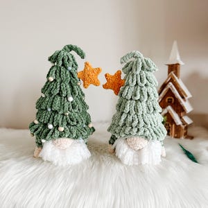 Könnte beinhalten: Zwei handgefertigte Weihnachtsbaum-Zwerge. Die Zwerge haben grüne, baumförmige Körper, weiße Bärte und beige Nasen. Jeder Zwerg hält einen orangefarbenen Stern. Die Zwerge sind mit kleinen weißen Perlen verziert. Im Hintergrund befindet sich eine kleine Holzkirche.