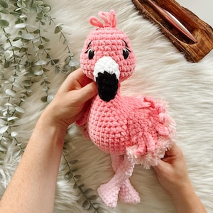 May include: A handmade pink crochet flamingo plush toy with a black beak, white accents, and black eyes. The flamingo has a fluffy texture and is held against a white, fluffy surface. The toy is a whimsical, handcrafted item.