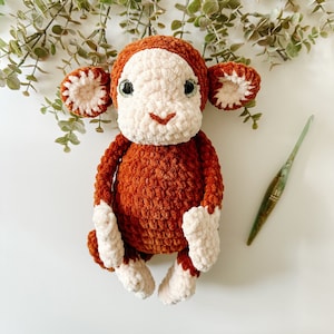 Może przedstawiać: Pluszowa zabawka małpki wykonana na szydełku, z brązowym futerkiem i białą buzią. Małpka ma duże, zielone oczy i mały, wyszyty uśmiech. Zabawka leży na białym tle.