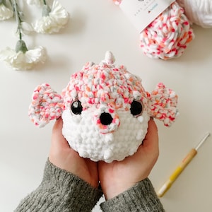 May include: A crocheted pufferfish toy in white and multi-coloured yarn, with large black eyes and orange details. The toy is held in hands, with yarn and a crochet hook visible. The yarn is labelled "Baby Snuggle" and "Honey Bunny".