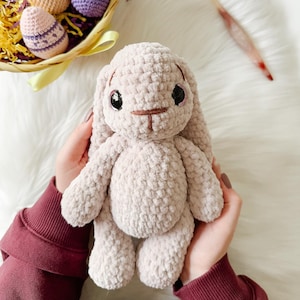 May include: A crocheted plush bunny toy in a light beige color, with embroidered eyes and a brown nose. The bunny is being held by a person's hands.
