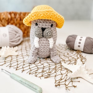 May include: A crocheted gray walrus wearing a yellow crocheted hat. The walrus is sitting on a brown fishing net with a white starfish and a crochet hook.