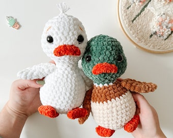 May include: Two crocheted stuffed duck toys. One duck is white with orange beak and feet. The other duck is green with brown and white accents and orange beak and feet.