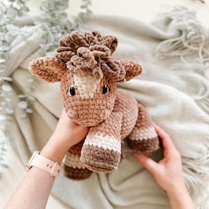 May include: A handmade crochet horse toy in shades of brown, tan, and white. The plush toy features a detailed mane, ears, and hooves. The horse is held up by hands, showcasing the soft texture and craftsmanship of the item.