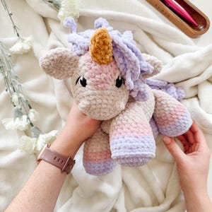 Può includere: Un giocattolo unicorno all'uncinetto fatto a mano in morbidi colori pastello. L'unicorno ha un corpo beige, un corno dorato e una criniera e coda lavanda. Il giocattolo è tenuto tra le mani su uno sfondo bianco e soffice.