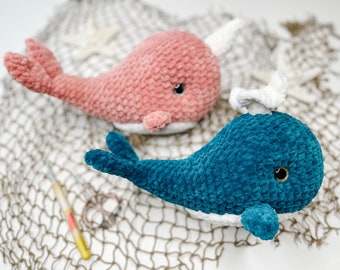 May include: Two crocheted whale plushies, one pink and one blue, on a white and brown net background.