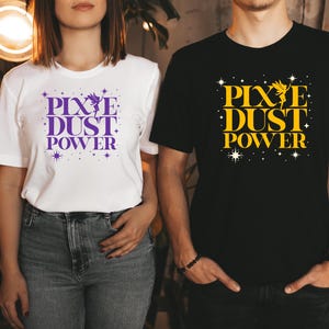 Puede incluir: Dos personas con camisetas a juego. Una camiseta blanca con texto morado "Pixie Dust Power" con estrellas. La otra camiseta es negra con texto amarillo "Pixie Dust Power" con estrellas.