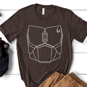 May include: A brown t-shirt with a white geometric design on the front. The design resembles a chest piece of armor with a crystal in the center. A small crescent moon symbol is on the right side of the design.