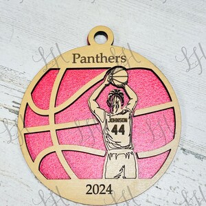 May include: Wooden basketball ornament with a pink background and a basketball design. The ornament features a basketball player in a jersey with the name "Johnson" and the number "44". The word "Panthers" is at the top, and "2024" is at the bottom.