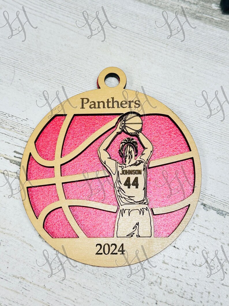 May include: Wooden basketball ornament with a pink background and a basketball design. The ornament features a basketball player in a jersey with the name "Johnson" and the number "44". The word "Panthers" is at the top, and "2024" is at the bottom.