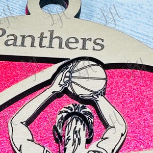 May include: A basketball-themed pendant with the word "Panthers" at the top. The pendant features a player in a jersey with the name "Johnson" on it, shooting a basketball. The background is pink.