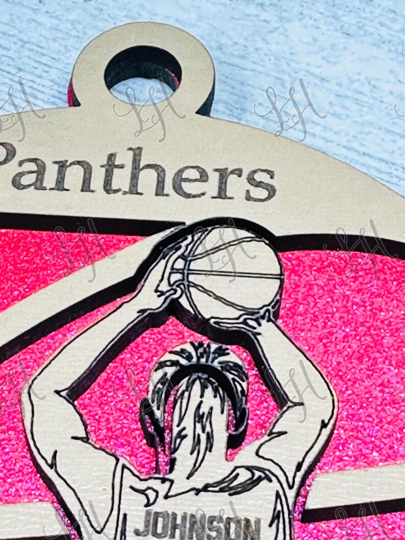 May include: A basketball-themed pendant with the word "Panthers" at the top. The pendant features a player in a jersey with the name "Johnson" on it, shooting a basketball. The background is pink.