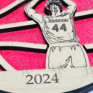 May include: Wooden basketball-themed ornament featuring a player in a jersey with "JOHNSON 44" on the back, shooting a basketball. The background is pink with black stripes, and the year "2024" is at the bottom. The ornament has a light brown wood finish.