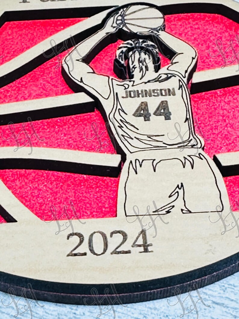 May include: Wooden basketball-themed ornament featuring a player in a jersey with "JOHNSON 44" on the back, shooting a basketball. The background is pink with black stripes, and the year "2024" is at the bottom. The ornament has a light brown wood finish.
