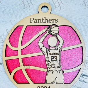 May include: A wooden basketball ornament with a pink background. The ornament features a basketball player shooting a ball, the word "Panthers", the year "2024", and the name "Watkins" with the number "23".