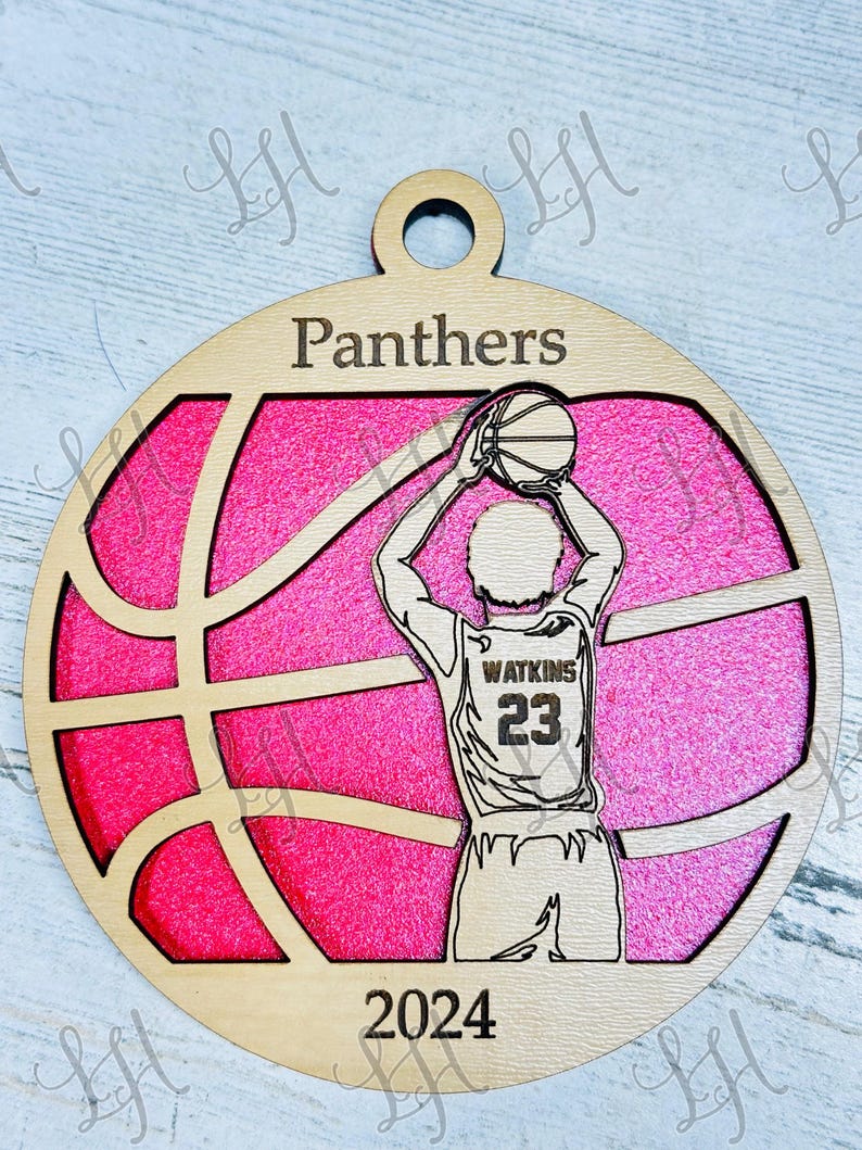 May include: A wooden basketball ornament with a pink background. The ornament features a basketball player shooting a ball, the word "Panthers", the year "2024", and the name "Watkins" with the number "23".