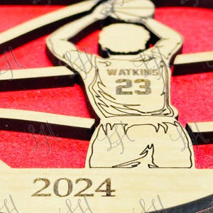 May include: Wooden basketball ornament featuring a player in a jersey with the name "Watkins" and the number "23". The ornament has a red background and the year "2024" engraved at the bottom. The word "Panthers" is at the top.