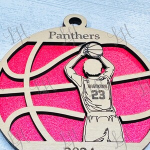 May include: Wooden basketball ornament featuring a basketball player in a shooting pose. The ornament has a pink background with a basketball design. The word "Panthers" is at the top, "Watkins 23" on the jersey, and "2024" at the bottom.