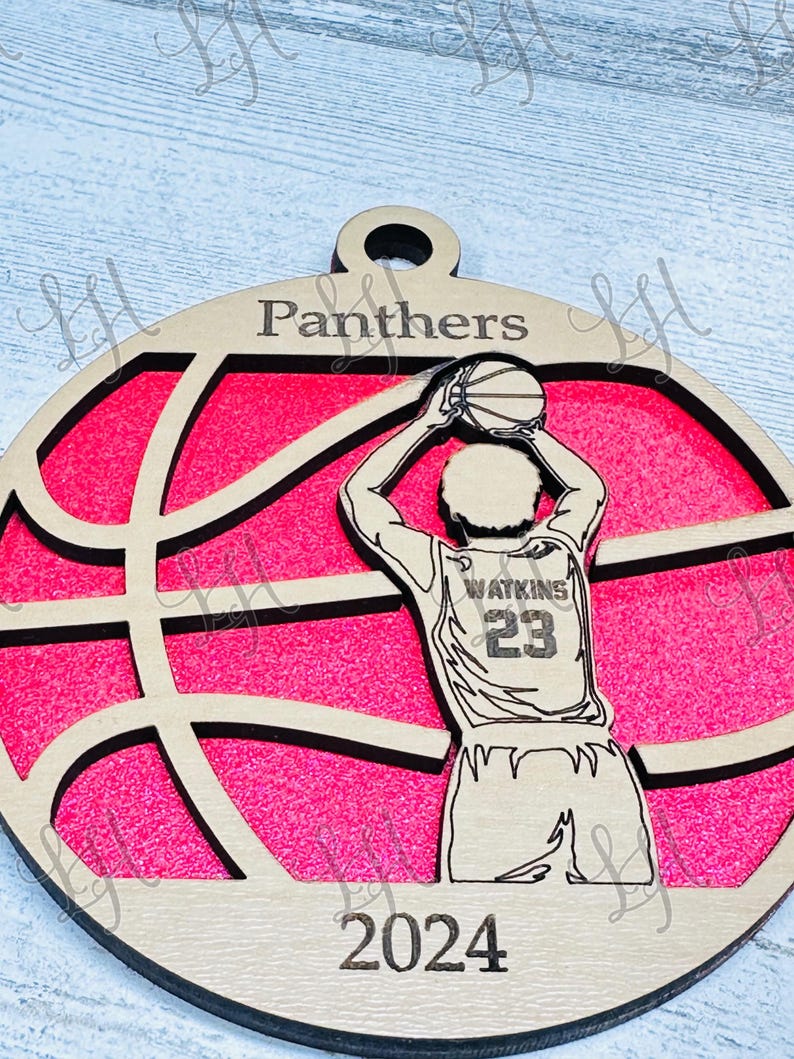May include: Wooden basketball ornament featuring a basketball player in a shooting pose. The ornament has a pink background with a basketball design. The word "Panthers" is at the top, "Watkins 23" on the jersey, and "2024" at the bottom.