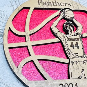 May include: Wooden basketball ornament with a pink glitter background. The ornament features a basketball player in a jersey with the name "Johnson" and the number "44". The word "Panthers" is at the top, and the year "2024" is at the bottom.