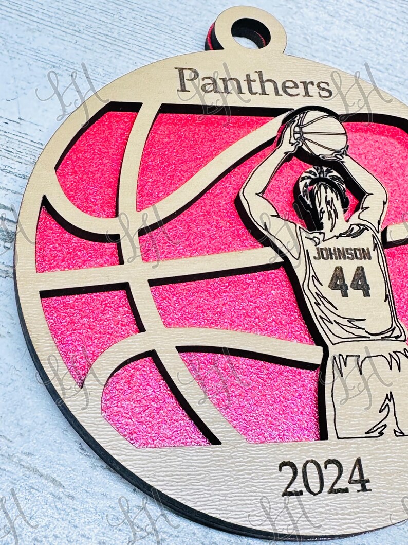 May include: Wooden basketball ornament with a pink glitter background. The ornament features a basketball player in a jersey with the name "Johnson" and the number "44". The word "Panthers" is at the top, and the year "2024" is at the bottom.