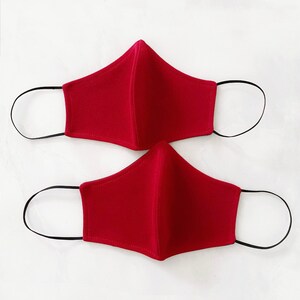 Pode incluir: Duas m&aacute;scaras de tecido vermelho com la&ccedil;os el&aacute;sticos pretos para as orelhas.