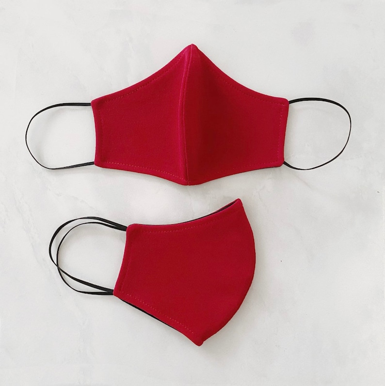 Pode incluir: Duas m&aacute;scaras de tecido vermelho com la&ccedil;os el&aacute;sticos pretos para as orelhas. As m&aacute;scaras s&atilde;o feitas de um material macio e respir&aacute;vel.