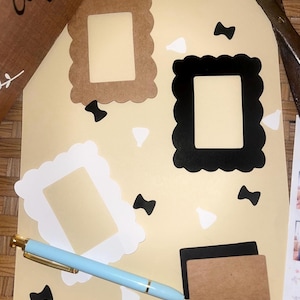 Puede incluir: Un conjunto de materiales de manualidades que incluye tres marcos decorativos en marrón, negro y blanco, con bordes festoneados. También hay un bolígrafo azul claro con detalles dorados y pequeños recortes de papel blanco y negro.