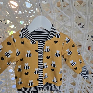 Könnte beinhalten: Senfgelbe Babyjacke mit Bienen- und Herzmuster. Die Jacke hat schwarz-weiß gestreifte Bündchen, Kragen und Saum. Die Jacke ist zugeknöpft und hängt an einem weißen Kleiderbügel.