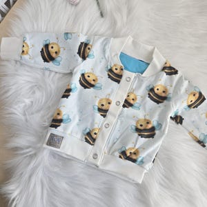 Könnte beinhalten: Eine weiße Babyjacke mit blauem Futter und einem Muster aus Comic-Bienen. Die Jacke hat einen Reißverschluss und Druckknöpfe.
