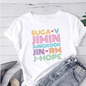 May include: A white t-shirt featuring the names of BTS band members in a vibrant, stacked design. The names, including SUGA, JIMIN, JUNGKOOK, JIN, RM, and J-HOPE, are in various colours. The shirt is styled with denim shorts and grey heels.