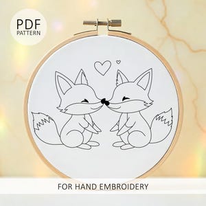 May include: A white embroidery hoop with a black and white fox design. Two foxes are kissing, with a heart above them. The text "PDF PATTERN" and "FOR HAND EMBROIDERY" are also visible.