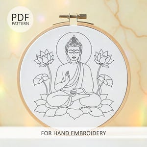 Pode incluir: Um bastidor de bordado com um desenho em tecido branco. O desenho é um desenho de linha preta de Buda em posição de lótus, com flores de lótus. O texto diz "PDF PATTERN" e "FOR HAND EMBROIDERY".