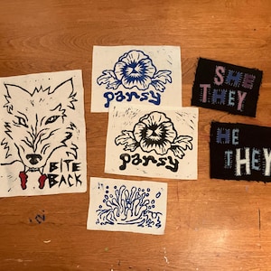 Może przedstawiać: Kolekcja ręcznie robionych naszywek i nadruków. Nadruki przedstawiają wilka z napisem "BITE BACK", niebiesko-biały kwiatowy wzór ze słowem "pansy" i abstrakcyjny niebieski wzór. Naszywki mówią "SHE THEY" i "HE THEY".