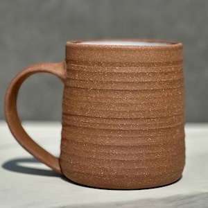 May include: A cylindrical ceramic mug with a speckled terracotta finish and a series of horizontal ridges encircling the exterior. The mug features a curved handle on the left side and a smooth, white glazed interior rim visible at the top opening.