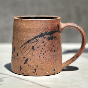 May include: A handmade ceramic mug with a speckled, reddish-brown exterior and a black interior. The mug features a curved handle and is decorated with black paint splatters. The mug is suitable for coffee, tea, or other beverages.