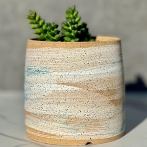 May include: A ceramic planter with a speckled, layered design in beige, white, and blue tones. The cylindrical pot holds a green succulent plant. The rim and base of the planter are a solid beige color.