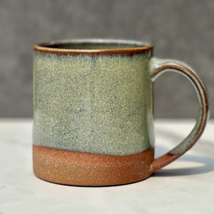 May include: A ceramic mug featuring a two-tone design with a speckled sage green glaze on the upper portion and a textured, unglazed reddish-brown clay base. The mug has a cylindrical body, a sturdy handle, and a smooth, rounded rim.