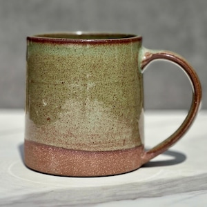 May include: A ceramic mug featuring a speckled sage green glaze on the upper portion and a solid reddish-brown base. The mug has a cylindrical body, a smooth rim, and a curved handle on the right side, showcasing a two-tone finish.