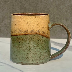 May include: A ceramic mug with a speckled finish, featuring a two-tone design with a light tan upper section and a sage green lower section. A horizontal band of small, dark brown bicycle illustrations encircles the middle of the mug.