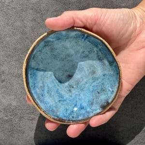 May include: A small, round ceramic dish featuring a mottled blue glaze with dark speckles and a textured, unglazed tan rim. The interior surface of the bowl displays variations in blue tones, ranging from light sky blue to deep navy, with a glossy finish.