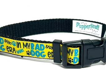 May include: Black dog collar with a yellow band featuring the text "MY BAD DOG ERA" in blue and paw prints. The collar has a black plastic buckle and a metal D-ring. The brand "PupperStuff" is visible.