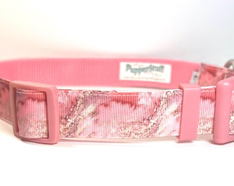 May include: Pink dog collar with a patterned ribbon design. The collar features a pink plastic buckle and a metal D-ring for leash attachment. The ribbon has a pink and white camouflage-like pattern. The brand name "Puppercraft" is visible on a label.