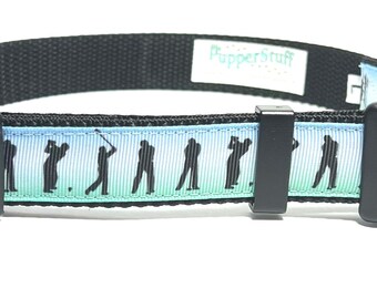 May include: Black dog collar with a golf theme. The collar features a black buckle and a patterned ribbon with silhouettes of golfers in action against a blue and green gradient background. The brand name "PupperStuff" is printed on a white label.