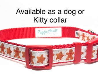 May include: Red dog or cat collar with a white band featuring gingerbread men and stars. The collar has a red plastic buckle and the text "Available as a dog or Kitty collar". The brand name "PupperStuff" is visible.