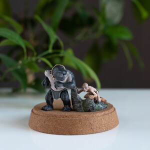 May include: A black and gray painted figurine of a creature from the Lord of the Rings movie series. The creature is holding a smaller figurine of a hobbit. The figurines are on a brown base.