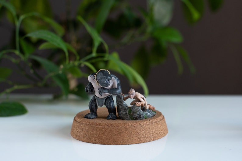 May include: A black and gray painted figurine of a creature from the Lord of the Rings movie series. The creature is holding a smaller figurine of a hobbit. The figurines are on a brown base.