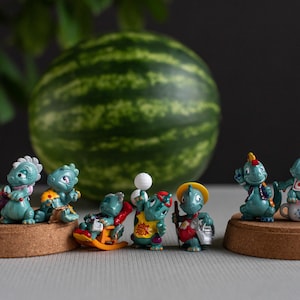 Könnte beinhalten: Eine Sammlung von acht kleinen türkisfarbenen Dinosaurierfiguren in verschiedenen Posen und Outfits, darunter eine im Schaukelstuhl, eine mit gelbem Hut und eine auf einer Tasse mit der Aufschrift Baby, angeordnet auf zwei runden Korksockeln.