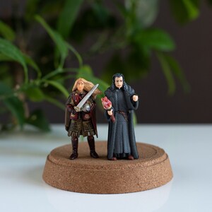 May include: Two miniature figurines on a cork base. One figurine is a woman with long blonde hair wearing a red and gold outfit and holding a sword. The other figurine is a man with black hair wearing a black robe and holding a red object.